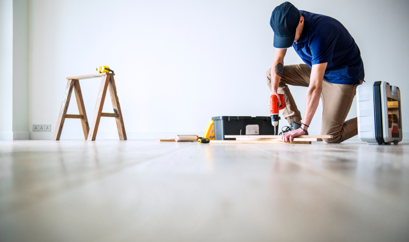 Tradesperson kneeling on a light wood floor using a cordless drill to install engineered hardwood flooring planks by Renovation King in Edmonton.